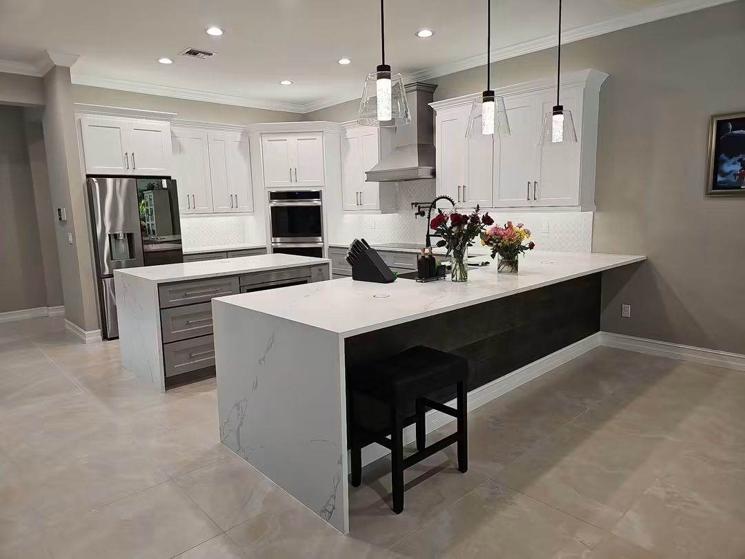 Same kitchen after remodel with two quartz waterfall islands, white shaker cabinets and wall ovens