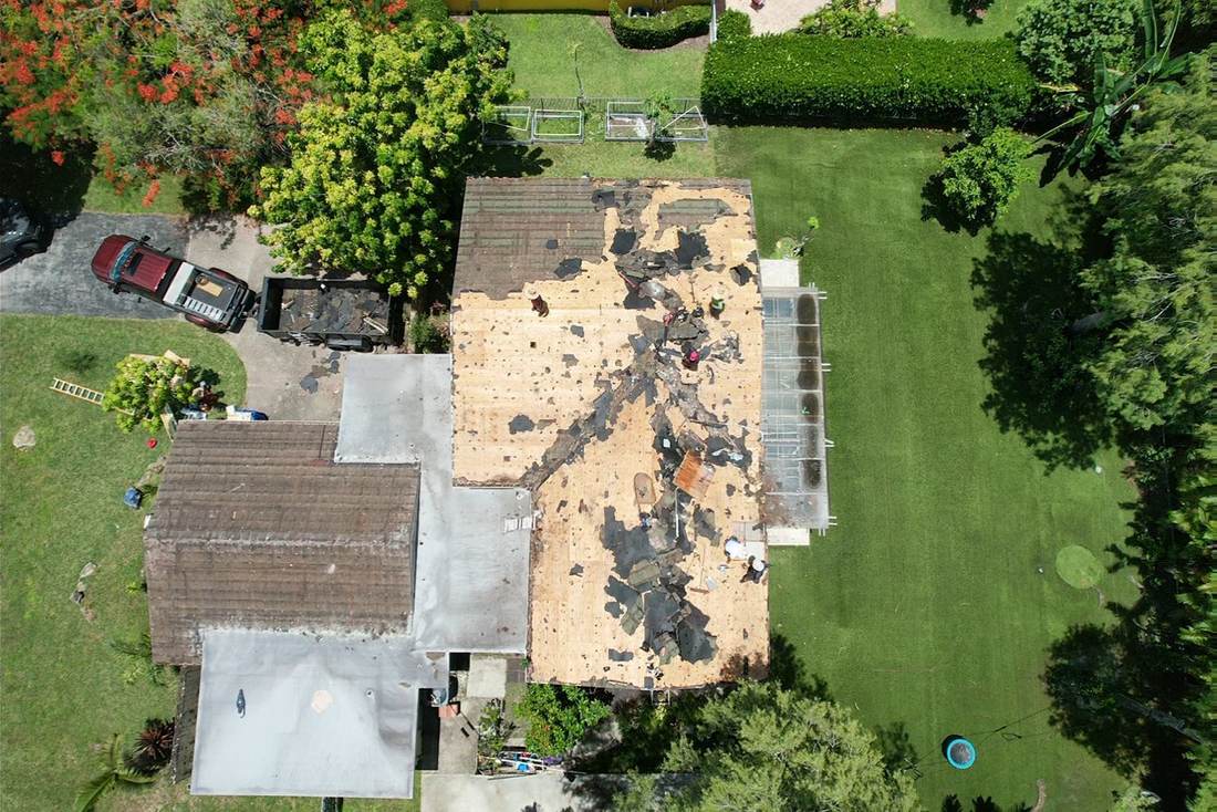 Aerial view of a full roof tear-off with crew working on the exposed deck and a dump trailer in the driveway